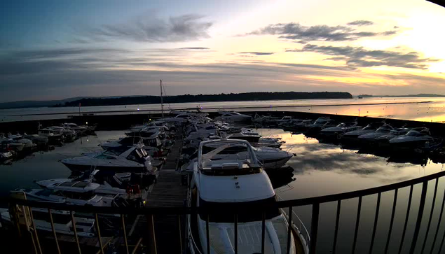 A marina filled with various boats is surrounded by calm water reflecting the sunset. The sky is filled with soft clouds and hues of orange and blue. In the foreground, there's a railing, and the boats are docked along a wooden pier. The scene conveys a peaceful evening atmosphere by the water.