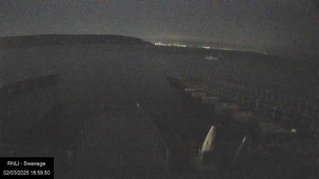 A dimly lit view of a harbor at night, with dark water reflecting faint lights in the distance. The silhouette of a pier can be seen, leading to a few boats moored along the edges. The shoreline is visible with a low hill in the background, while the sky above is mostly dark, suggesting an overcast evening.