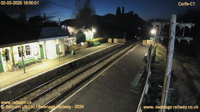 A dimly lit train station platform is visible, featuring a stone building with large windows, a green bench, and a sign that reads "WAY OUT." The platform is empty, with two railway tracks running parallel. In the background, there are chairs and a wooden fence, and a telephone pole with wires on the right side. The sky is darkening with shades of blue, indicating evening time.