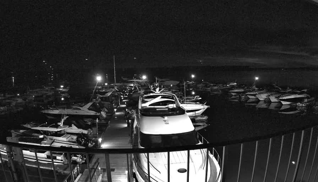 A nighttime scene of a marina filled with various boats and yachts. Some boats are docked along a wooden walkway that leads into the water. The scene is illuminated by several bright lights, reflecting softly on the calm water surface. The sky is dark with hints of clouds, and the overall ambiance is tranquil and serene.