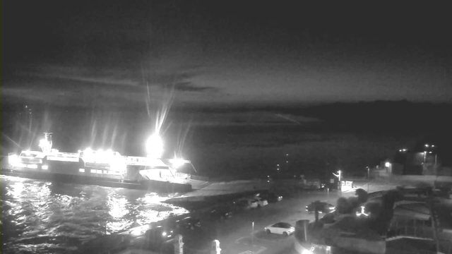 A night scene featuring a ferry docked at a pier, illuminated by bright lights. The water reflects these lights, creating shimmering patterns. In the foreground, there are parked cars, and a dimly lit area with some structures, possibly a building or cafe, visible on the right side. The sky is dark with faint clouds, and the atmosphere is calm and quiet.