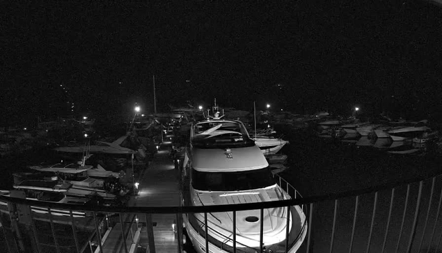 A nighttime view of a marina filled with boats. In the foreground, a large white yacht is docked, illuminated by nearby lights. Surrounding it are various smaller boats, some covered with tarps, and others parked along wooden docks. The scene is predominantly dark, with scattered lights reflecting on the water, creating a tranquil ambiance.