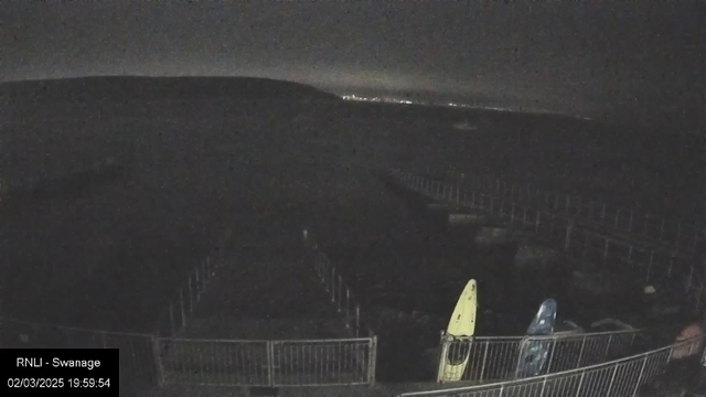 A dimly lit scene shows a coastline with a faint outline of hills in the background. In the foreground, there are two kayaks resting against a railing, one yellow and one blue. The area appears to be empty and quiet, with no visible people. The water extends into the darkness beyond the shoreline, and city lights can be faintly seen in the distance under a night sky.