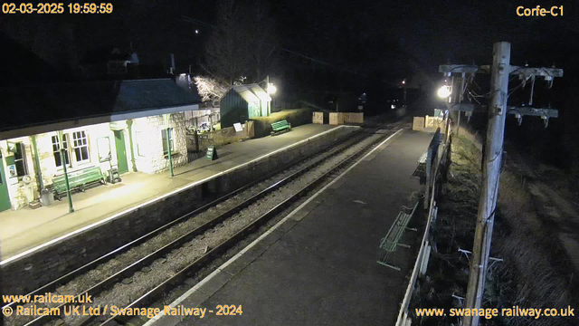 A dimly lit railway station at night. The scene shows two railway tracks running parallel, with a few benches along the platform. On the left side, there is a small stone building with large windows, and a green bench is positioned nearby. A green wooden structure is also visible in the background. A wooden fence marks the end of the platform on the right. A street lamp and utility pole are present, providing some light in the area. The sky is dark, indicating it is nighttime.