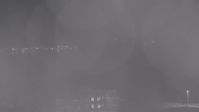 A dark, nighttime view captured from a webcam. The foreground features a dimly lit multi-story building with some windows showing faint light. In the background, there is a body of water reflecting vague lights from distant sources, likely the shoreline or buildings across the water. The overall scene appears hazy and lacks detail due to low visibility.