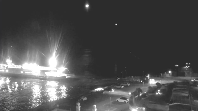 A nighttime scene showing a ferry with bright lights docked at a port, casting a reflection on the calm water. In the foreground, there are several parked vehicles, illuminated by streetlights. The background features a dark sky, partially obscured by a few distant stars, and a faint hint of the shoreline.