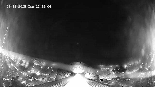 Black and white image from a webcam showing a low-light urban scene. The bottom of the image features buildings and roads, with faint outlines of structures illuminated by surrounding lights. The top half of the image is mostly dark, creating a stark contrast with the bright light emanating from the center, suggesting an open space or exit. A timestamp in the upper left corner reads "02-03-2025 Sun 20:01:04," and the lower left corner includes the text "Powered By Brighton CCTV."