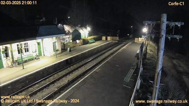 A dimly lit railway station platform at night. The scene features a stone building with a green roof and several windows, partially obscured by darkness. There are a couple of green benches on the platform, and a sign that reads "Way Out" is visible. The railway track runs parallel to the platform, and a wooden fence is seen in the background. A utility pole is on the right side of the image, with wires and a light fixture. The overall atmosphere is quiet and desolate, typical of a nighttime setting.