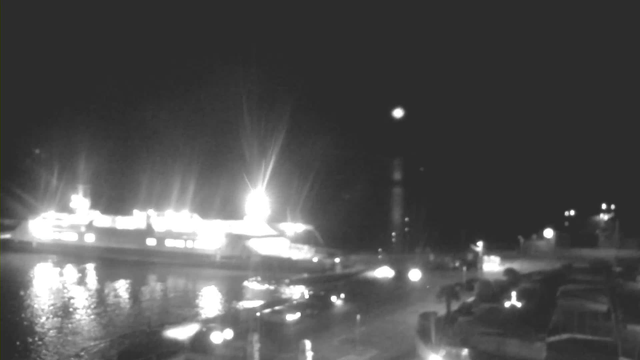 A night scene at a waterfront. In the foreground, several dark shapes appear to be vehicles parked along the edge. The water reflects shimmering lights from a nearby ferry that is brightly illuminated, creating a contrast against the dark surroundings. The ferry is docked, with some equipment visible on its deck. In the background, there are faint lights from buildings or streetlamps, creating a soft glow against the night sky.