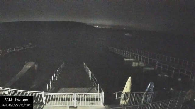 A dark scene featuring a waterfront area with a wooden dock extending into the water. The shoreline and hills are visible in the background, blending into the night sky. Two canoes, one yellow and one blue, are positioned on a platform at the edge. The image is dimly lit, indicating nighttime.