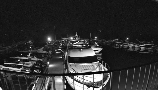 A view of a marina at night, with several boats docked along a wooden pier. The scene is predominantly dark, illuminated by scattered lights on the boats and along the dock. A large white boat is positioned prominently in the foreground, with smaller boats visible in the background. The reflection of the lights sparkles on the water surface. A railing is visible in the foreground, framing the scene.