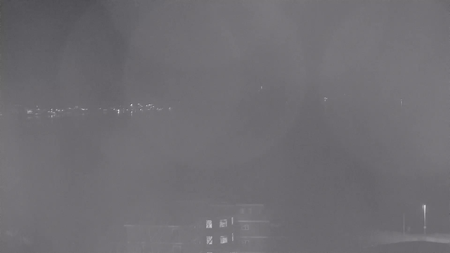 A dimly lit scene captured from a webcam showing a distant city skyline. The view is mostly obscured by fog, with faint outlines of buildings and a few scattered lights visible in the background. The foreground features an indistinct structure, possibly an incomplete silhouette of a building, with some illuminated windows visible, while the sky appears overcast and dark.