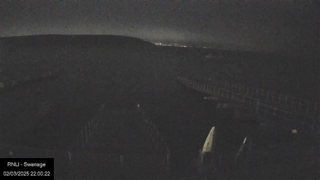 A dark, nighttime scene of a coastal area with silhouettes of boats and a wooden pier. In the background, hills are visible under a slightly illuminated sky with distant lights suggesting a town or city. The overall atmosphere is calm and quiet, with minimal visible detail due to low light.