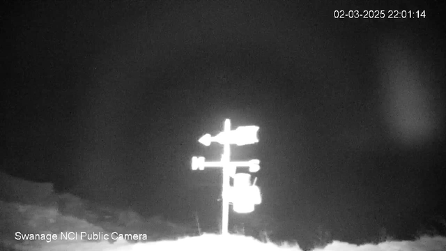 The image shows a black-and-white infrared view of a weather vane, positioned on a pole, with directional indicators for north, south, east, and west. The weather vane has an arrow pointing to the left, indicating wind direction. At the bottom corner of the image, there is a timestamp displaying "02-03-2025 22:01:14," and text indicating "Swanage NCI Public Camera." The background is dark with little visibility beyond the weather vane.