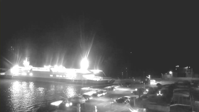 A brightly lit ferry docked by the water at night. The ferry has multiple lights on its side and the upper deck, illuminating the surrounding area. In the foreground, several parked cars are visible along the waterfront, with reflections on the water's surface. The background shows a few lights along the shoreline, indicating buildings or streetlights. The scene is primarily in shades of black and white, creating a high-contrast image.