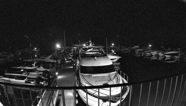 A nighttime image of a marina, featuring several boats docked in a harbor. A prominent white yacht is in the foreground, with a ramp leading to it. There are additional boats of various sizes and shapes visible in the background, with faint lights reflecting on the water. The scene is predominantly dark, with some illuminated areas creating contrast.