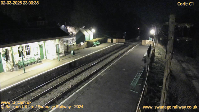 A dimly lit railway station platform at night. The foreground features two railway tracks running parallel to the viewer. To the left, there is a stone building with large windows and a green bench underneath a covered area. There is a sign that reads "NO EXIT" near the benches. The right side has a small wooden fence enclosing a green bench and some plants. The background includes trees and distant lights, hinting at a rural setting. The overall atmosphere is quiet and serene, illuminated by soft lighting.