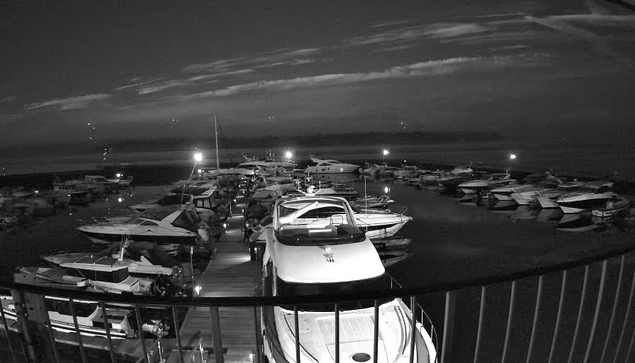 A nighttime view of a marina filled with various boats docked along a wooden pier. The scene is illuminated by artificial lights reflecting on the water's surface, highlighting numerous boats of different sizes and shapes. The sky is partially cloudy, with hints of light from distant sources. The atmosphere is calm and serene, typical of a twilight setting near the water.