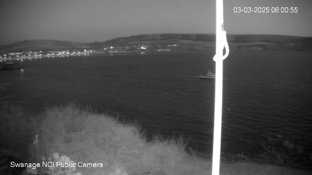 A black and white image showing a calm body of water with distant hills in the background. A small boat is visible on the water. The shoreline is lined with lights indicating buildings, creating a subtle glow along the coast. The scene is captured at dawn or dusk, with a slightly cloudy sky. A pole with a camera is positioned on the left side of the frame. There is a timestamp in the corner indicating the date and time.