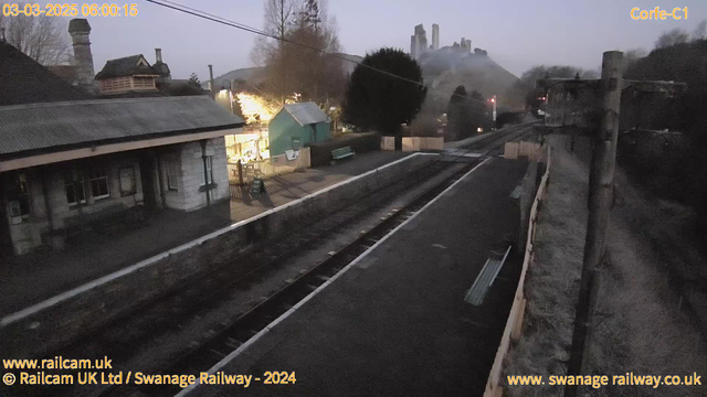 A dimly lit railway station platform is shown, with two sets of railway tracks running parallel. On the left, there is a stone building with a peaked roof, and a small outdoor area with a green wooden bench. Some trees are visible in the background, partially obscuring a distant hill with ruins. The right side of the image includes a wooden fence along the tracks and a utility pole. The sky is lightening, suggesting early morning, while a few lights are seen illuminating the area.