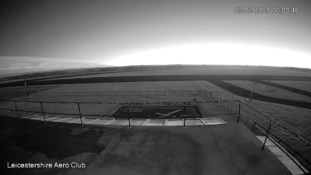 A black and white image captured from a webcam showing a wide view of an airfield at dawn. The foreground features a flat rooftop with a railing on the left side. Below the rooftop is a helipad marked with an "X". The background reveals a vast expanse of grass, with faint lines indicating runways. The sky is bright near the horizon, transitioning to a darker tone above as it approaches the top of the image. The date and time in the upper right corner indicate March 3, 2025, at 6:00:16 AM.