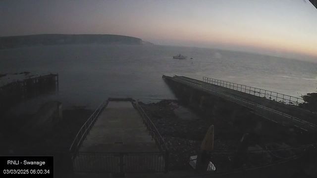 A serene early morning view of the sea, with a light fog covering the horizon. The water appears calm and slightly reflective, showing soft gradients of color as the sun rises. A small boat can be seen floating on the water. In the foreground, a jetty extends toward the water, partially obscured by rocks and debris, with a railing along its edge. To the right, a surfboard is leaned against the jetty. The scene is tranquil, evoking a sense of peace and the start of a new day.