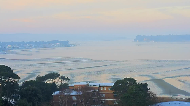 A serene view of a calm body of water during early morning light. The surface reflects soft pastel colors, primarily pale blues, pinks, and yellows. In the foreground, there are patches of trees and a building with a flat roof, partially covered in snow. In the distance, the shoreline features a small town with scattered buildings. The horizon is blurred, creating a misty effect that softens the landscape. Gentle ripples are visible in the water, adding a sense of tranquility to the scene.