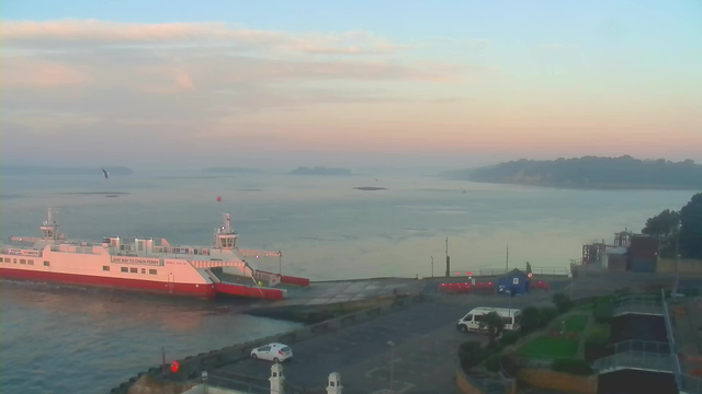 A large ferry boat is docked at a pier, with a low tide revealing the muddy shoreline. In the background, calm water stretches out towards a faint shoreline of hills under a pastel-colored sky during dawn or dusk. Several vehicles are parked near the pier, and greenery is present in the foreground.