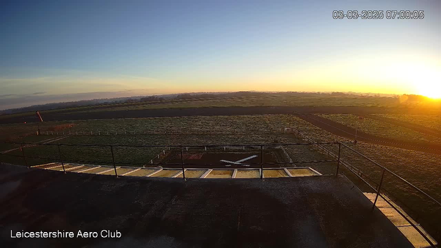 A panoramic view of a grassy field at sunrise, with a clear blue sky gradually changing to hues of orange and yellow near the horizon. In the foreground, there is a flat rooftop with a railing. Below the rooftop, there is a grassy runway marked with white lines forming a cross shape. The area is peaceful, with no visible aircraft or people, highlighting the dawn's quiet atmosphere.