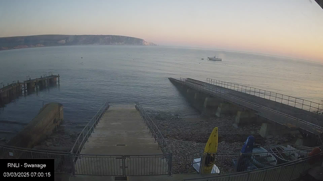 A calm sea scene at dawn, with gentle waves lapping at the shore. In the foreground, a ramp leads down to the water, flanked by rocky shores and a few boats tied up at a small pier to the left. To the right, another pier extends into the water with a railing alongside. A couple of colorful kayaks, yellow and blue, are visible on the right side. In the background, a rocky coastline fades into the horizon, where the sky transitions from light orange to pale blue as the sun rises.