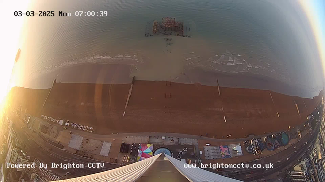 A view from a high vantage point overlooking a beach at sunrise. The sandy beach stretches across the bottom of the image, with a few wooden groynes visible in the sand. Gentle waves wash onto the shore. In the distance, partially submerged ruins of a pier can be seen in the water. Along the beach, there are structures, including colorful awnings and a circular feature, hinting at amusement or entertainment areas. The sky is brightening with shades of yellow and orange, indicating early morning light. The date and time in the corner indicate it is March 3, 2025, at 7:00 AM.