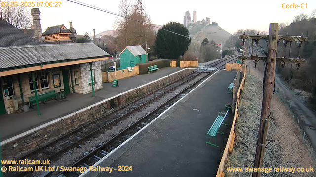 A view of a railway station in a rural area during early morning. The platform is made of dark stone, with a few green benches and a wooden fence. A small green building is visible on the left side, with a sign indicating "WAY OUT" nearby. In the background, a castle ruin sits atop a hill, surrounded by trees. The sky is lightening, suggesting dawn. The railway tracks run parallel to the platform, leading into the distance. There are utility poles with wires along the edge of the platform.