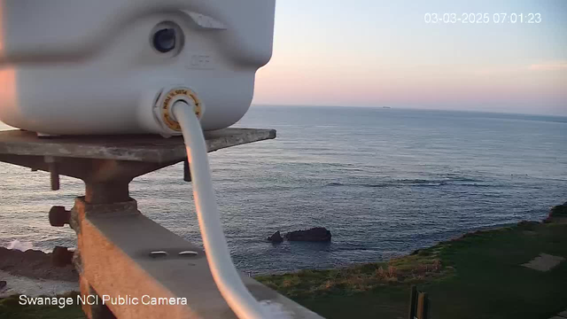 A close-up view of a white container mounted on a railing, with a hose extending from the bottom. In the background, the ocean is visible with gentle waves under a pastel-colored sky at dawn. A rocky formation is partially visible in the water, and the date and time are displayed in the top right corner. The scene is peaceful with no people present.