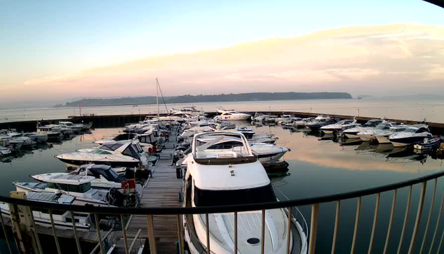 A scenic view of a marina at dawn. Numerous boats are anchored in calm waters, reflecting the soft colors of the sunrise. A wooden dock is visible in the foreground, with boats lined up on either side. The distant land shows a gentle slope and a hazy atmosphere, while a few fluffy clouds are visible in the light blue sky.