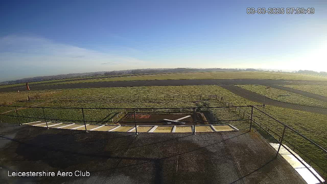 A wide view from a webcam showcases a clear blue sky above the Leicestershire Aero Club. In the foreground, a flat rooftop with a railing surrounds a landing area marked with an "X." To the right, a grassy field stretches into the distance, leading to a tarmac runway that curves gently. A distant hill and low vegetation are visible beyond the runway, indicating an open landscape under sunny conditions. The scene captures a serene and expansive outdoor setting.