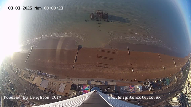 Aerial view of a beach with a sandy shoreline and gentle waves lapping at the water's edge. There are remnants of a pier visible partially submerged in the water. Various colorful structures and vehicles can be seen along the strand, with a road running parallel to the beach. The sky is clear, and the sunlight reflects off the water, creating a bright glare in the upper left corner. The timestamp in the corner indicates it's early morning on March 3, 2025.