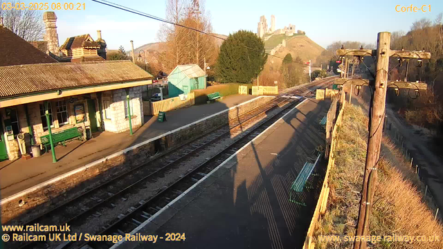 A railway station is depicted in the image, bathed in warm sunlight. The foreground features a platform with several green benches, accompanied by a stone wall at its edge. A traditional railway shelter with a sloped roof is visible, along with signs indicating exits. In the background, a hill rises with a castle or ruins atop, partially obscured by trees. The sky is clear, and a telephone pole with wires runs along the edge of the platform.