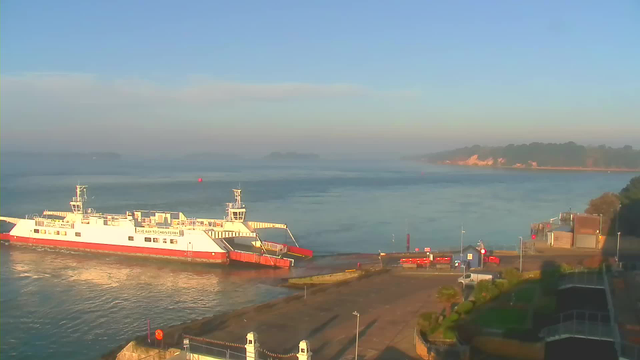 A view of a calm body of water with a ferry docked at a pier. The ferry is white with red accents and has a ramp lowered to the dock. In the background, there are green hills and a sandy coastline shrouded in light mist under a clear blue sky. On the pier, there are some vehicles and markers, with a few palm-like plants nearby. The scene is illuminated by the soft morning light.