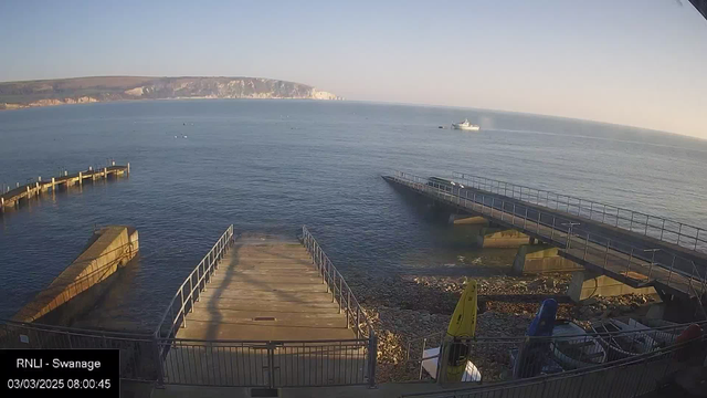 A view of a calm sea at sunrise, with a rocky coastline in the background. There are two piers extending into the water, one built of wooden beams and the other of concrete. Several boats can be seen on the water, and there are kayaks and other equipment stored on the shore. The sky is clear, displaying a gradient from light blue to soft gold as the sun rises.