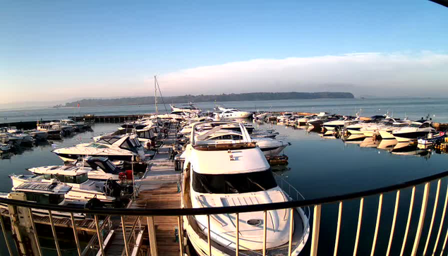 A tranquil marina scene with numerous boats docked at a wooden pier. The water is calm, reflecting the boats and the clear blue sky. In the background, a faint shoreline with trees and gentle hills is visible under a soft haze. The image captures a sense of serenity, with no people in sight, and the sunlight casts a warm glow over the scene.