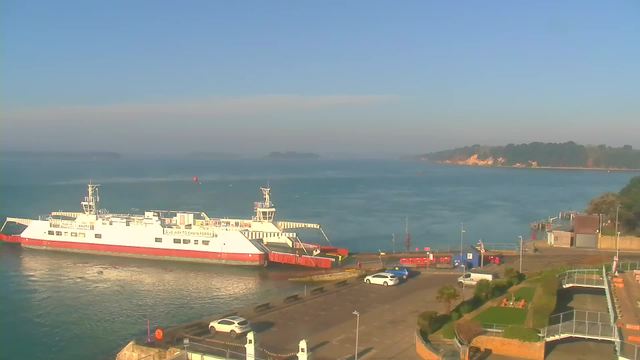 A white ferry with red accents is docked at a harbor, surrounded by calm blue water. There are several parked cars on a paved area, and to the right, there is a grassy area with chairs. In the background, there are trees and a rocky shore under a clear blue sky. The scene is bright and sunny, with a few distant islands visible beyond the ferry.