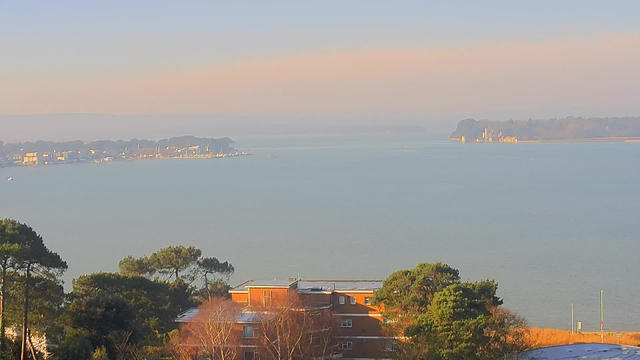 A scenic view of a calm body of water, likely a bay or harbor, under a soft blue sky. In the foreground, there are several green trees and a residential building with brown walls. To the left, boats can be seen scattered across the water, and in the distance, a shoreline lined with trees and buildings is visible. The atmosphere is serene and slightly misty, suggesting a peaceful morning with gentle light.