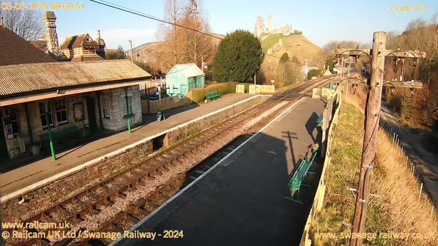 A railway station scene during daylight. The foreground shows a platform with stone edges, parallel railway tracks, and a wooden utility pole on the right. A green wooden bench and a small blue building are visible on the platform. In the background, a hill rises with castle ruins at the top, surrounded by trees. The sky is clear and blue.