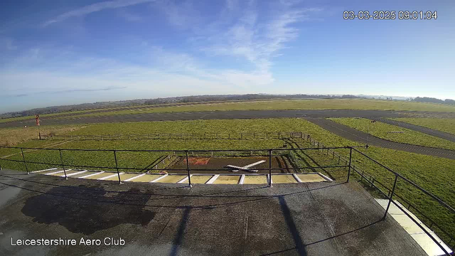 A view from a webcam mounted on a building, overlooking a grassy airfield. In the foreground, a helipad is marked with white and red paint. Beyond the helipad, there is an open field with some patches of grass. To the left, there is a windsock indicating wind direction. In the background, the airfield runway stretches out, bordered by low fences. The sky is mostly clear with a few clouds, and the image captures bright daylight. The date and time in the top right corner indicate March 3, 2025, at 09:01 AM.