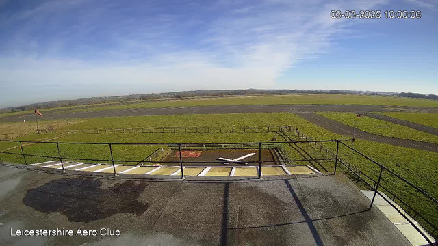 A clear view from a webcam at Leicestershire Aero Club shows a wide grassy airfield under a blue sky with scattered clouds. In the foreground, there is a helipad marked with a white cross, and the ground is wet, likely from a recent rain. A flag pole with an orange and white windsock indicates wind direction. In the background, the airfield extends into the distance with patches of grass and some distant trees on the horizon. The date and time is displayed in the upper right corner.