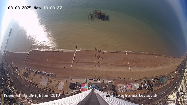 A wide-angle view from a high vantage point overlooking a sandy beach and the sea. The water is calm with sunlight reflecting off the surface. Several people are walking along the beach, and wooden structures are visible in the water, likely remnants of a pier. To the left, a pier extends out over the water. On the land, various colorful structures and attractions, such as a amusement park, are visible along the beachfront. The date and time are displayed in the upper left corner.