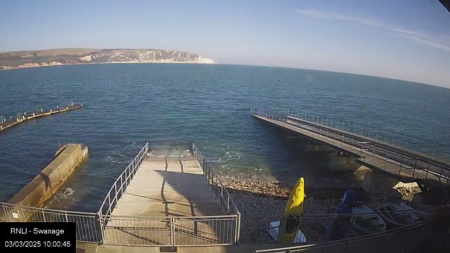 A view of the ocean with a clear blue sky above. In the foreground, there is a wooden ramp leading down toward the water, bordered by railings. To the right, a small section of rocky beach is visible, along with several boats and kayaks stored on the shore. In the background, a white cliff coastline stretches along the horizon, and rippling waves can be seen on the surface of the water. The image is timestamped at 10:00:46 on March 3, 2025.