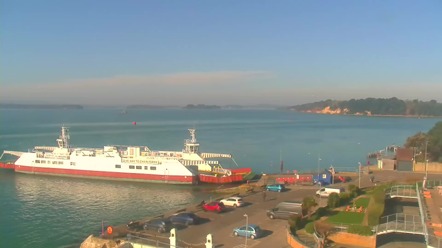 A large white ferry with a red underside is docked at a harbor. The water is calm and blue, reflecting the clear sky above. In the foreground, there are several parked cars of various colors, including red and blue. To the right, there is a small building and some greenery, while on the left, there are people moving around the dock area. The horizon features distant land with trees and cliffs, under a bright and sunny sky.