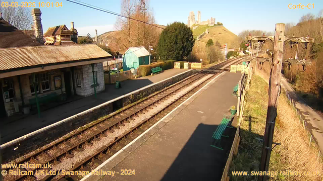 A sunny view of Corfe Castle railway station. The foreground features a platform with wooden benches in green and a sign indicating "WAY OUT." To the left, a stone building with a sloped roof and chimney is visible, suggesting it might be the ticket office. In the background, a hill with the ruins of Corfe Castle is set against a blue sky. Lush greenery surrounds the area, and there are power lines running overhead. The tracks run parallel to the platform, leading into the distance.