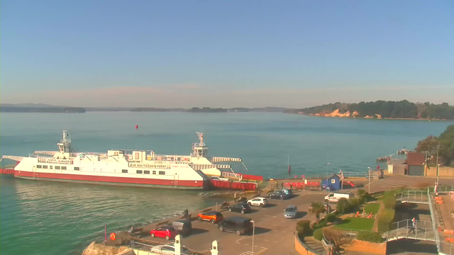 A ferry is docked at a waterfront terminal, with calm blue waters surrounding it. Nearby, several parked cars are visible along the shore, including a bright orange car. In the background, there are trees and a gently sloping coastline, with hills in the distance under a clear blue sky. A small pier extends into the water, and there are a few people near the ferry terminal.